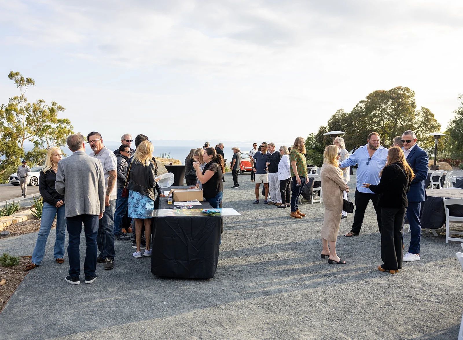 People gathered outdoors at a social event or reception near a scenic view with trees, cars parked in the background, and tables set up for guests.