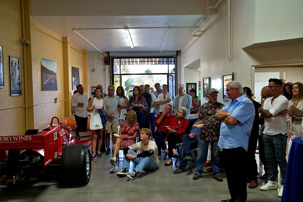 A group of people watching a red Formula 1 car on display in an indoor setting, with some seated and others standing.