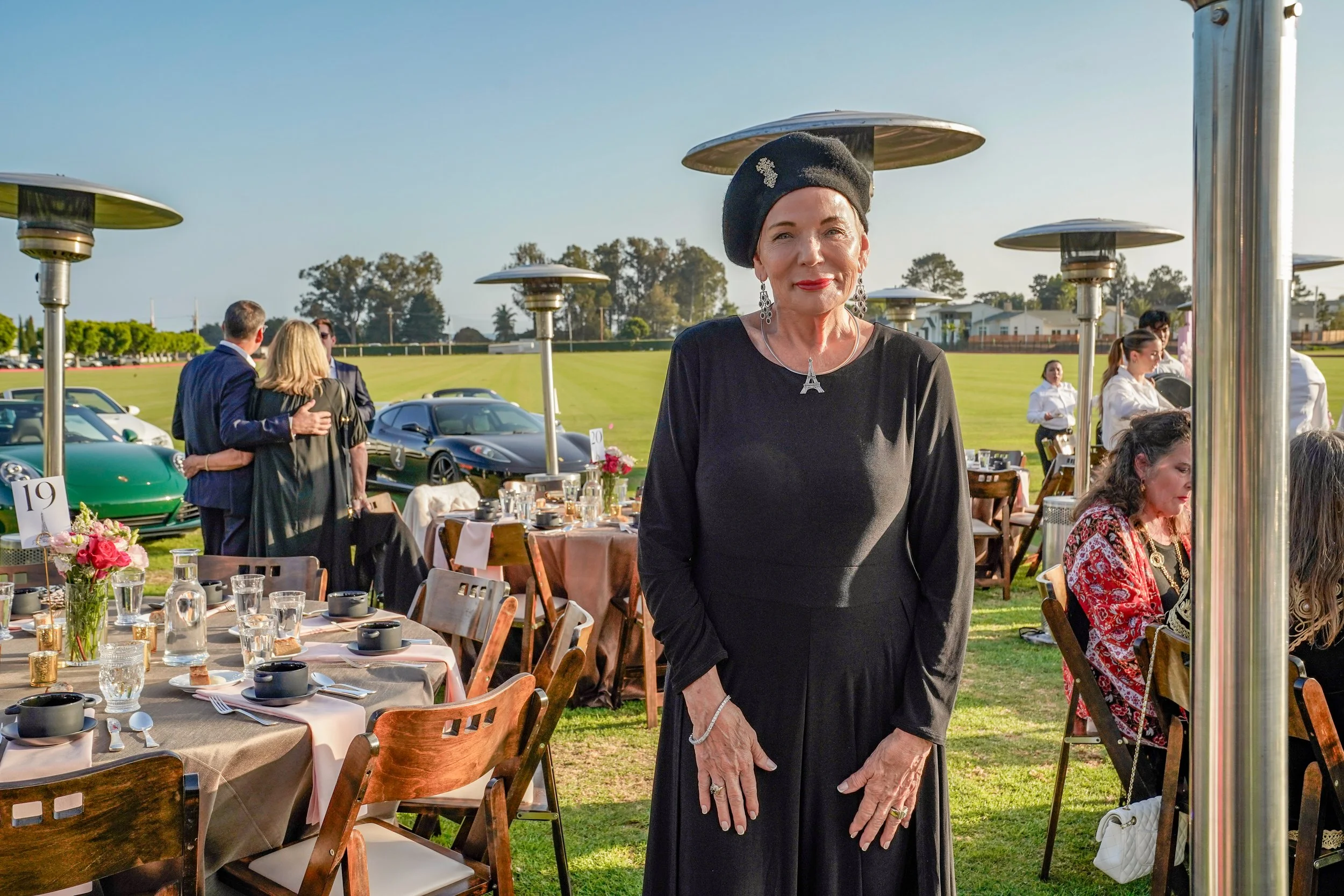 A woman dressed in black standing at an outdoor event with tables, chairs, and table settings, with other guests and vintage cars in the background. The event is held in a grassy area on a clear day.