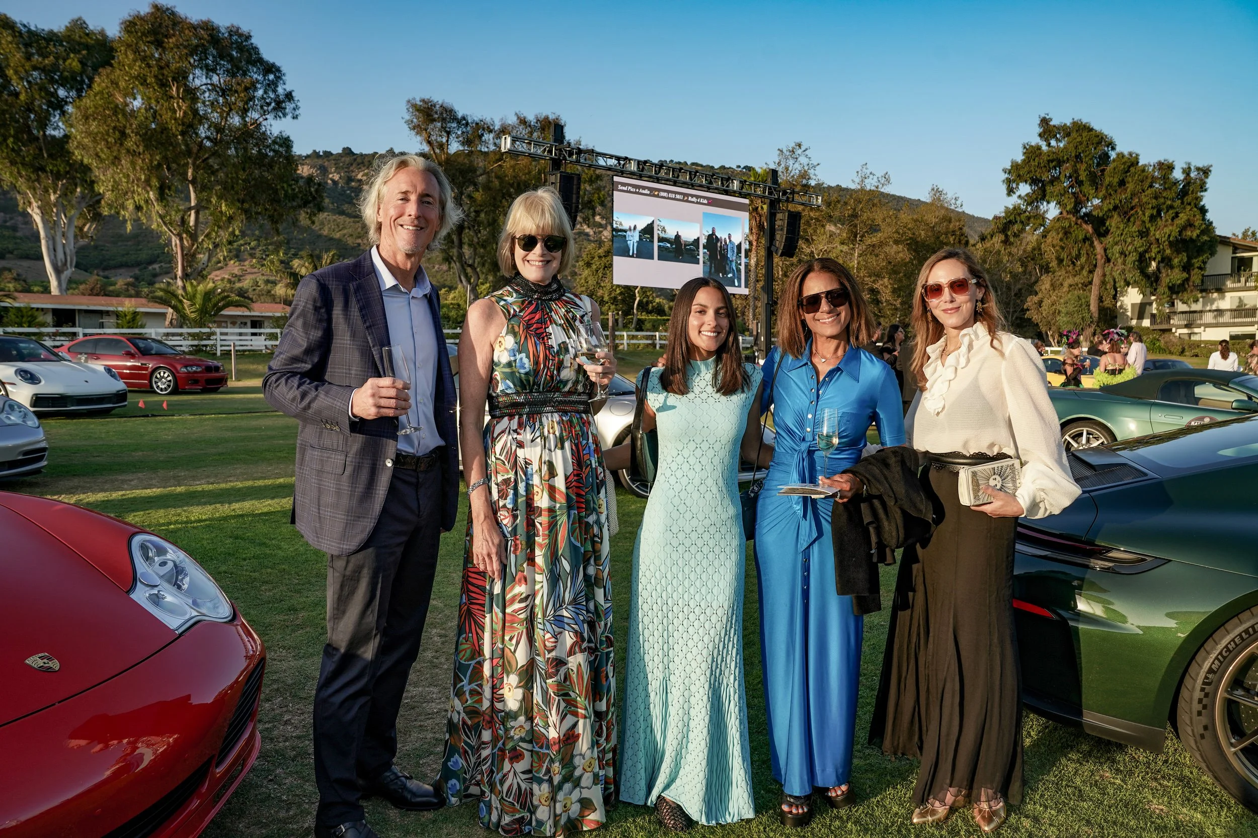 Group of five people standing outdoors at a car event, dressed in formal and fashionable attire, with luxury sports cars and a large screen in the background.