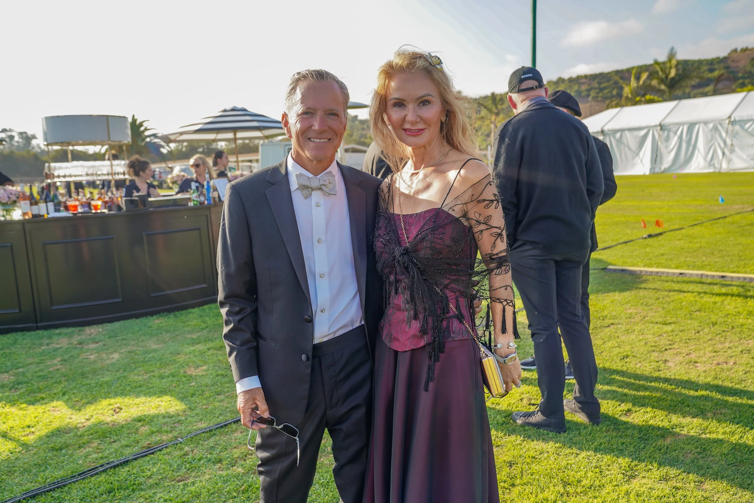 A man in a tuxedo and a woman in an elegant dress standing outdoors at a formal event, smiling at the camera, with a grassy area and other people in the background.