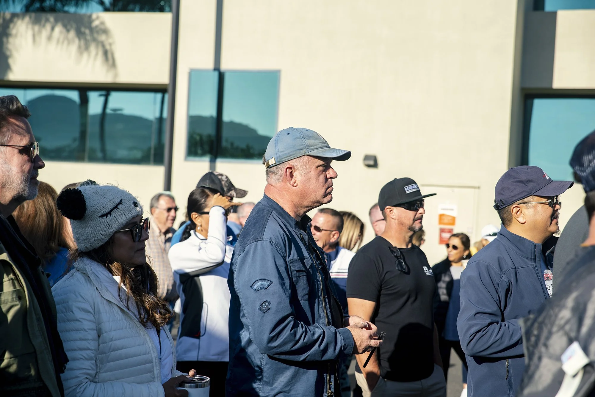 A group of people gathered outside near a building, some wearing sunglasses and hats, with a mountain and glass windows in the background.