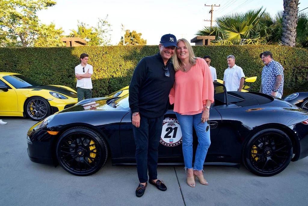 A man and woman stand together in front of a black sports car at a car gathering, with other luxury cars and people in the background.