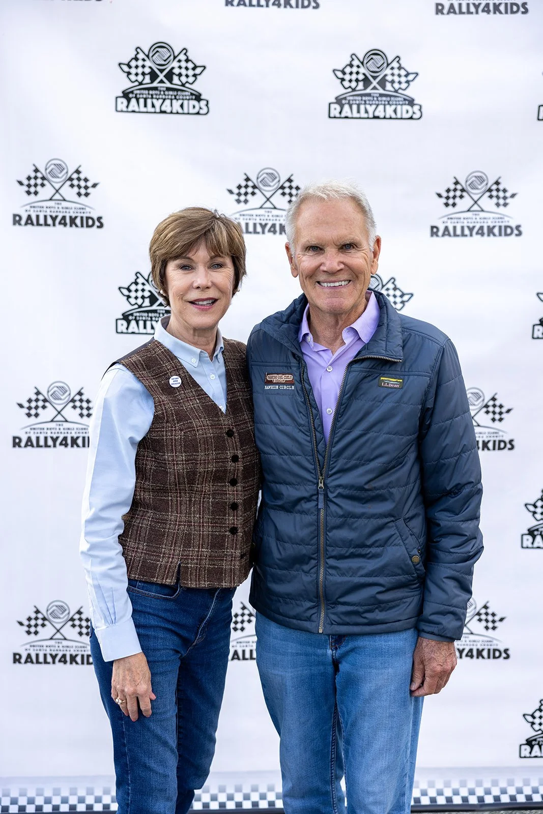 A woman and a man standing together smiling in front of a Rally4Kids backdrop. The woman is wearing a brown plaid vest over a light blue button-up shirt and blue jeans. The man is wearing a blue quilted jacket over a light purple shirt and blue jeans