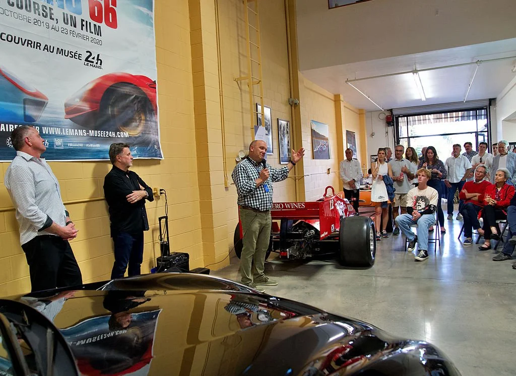 A man speaking into a microphone in front of an audience, with a race car and a large poster featuring a red sports car in the background inside a building.