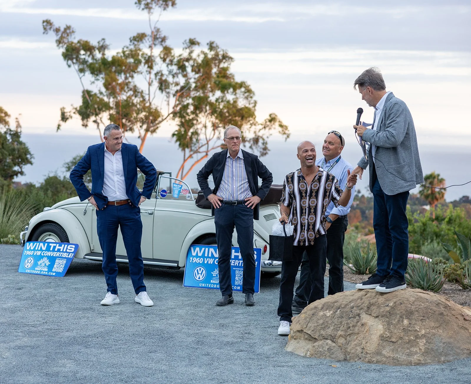 A group of five men at an outdoor event, with a vintage white Volkswagen convertible car and signs promoting a giveaway of a 1960 VW convertible. One man on the right is holding a microphone and shaking hands with another man on a rock, all smiling. 