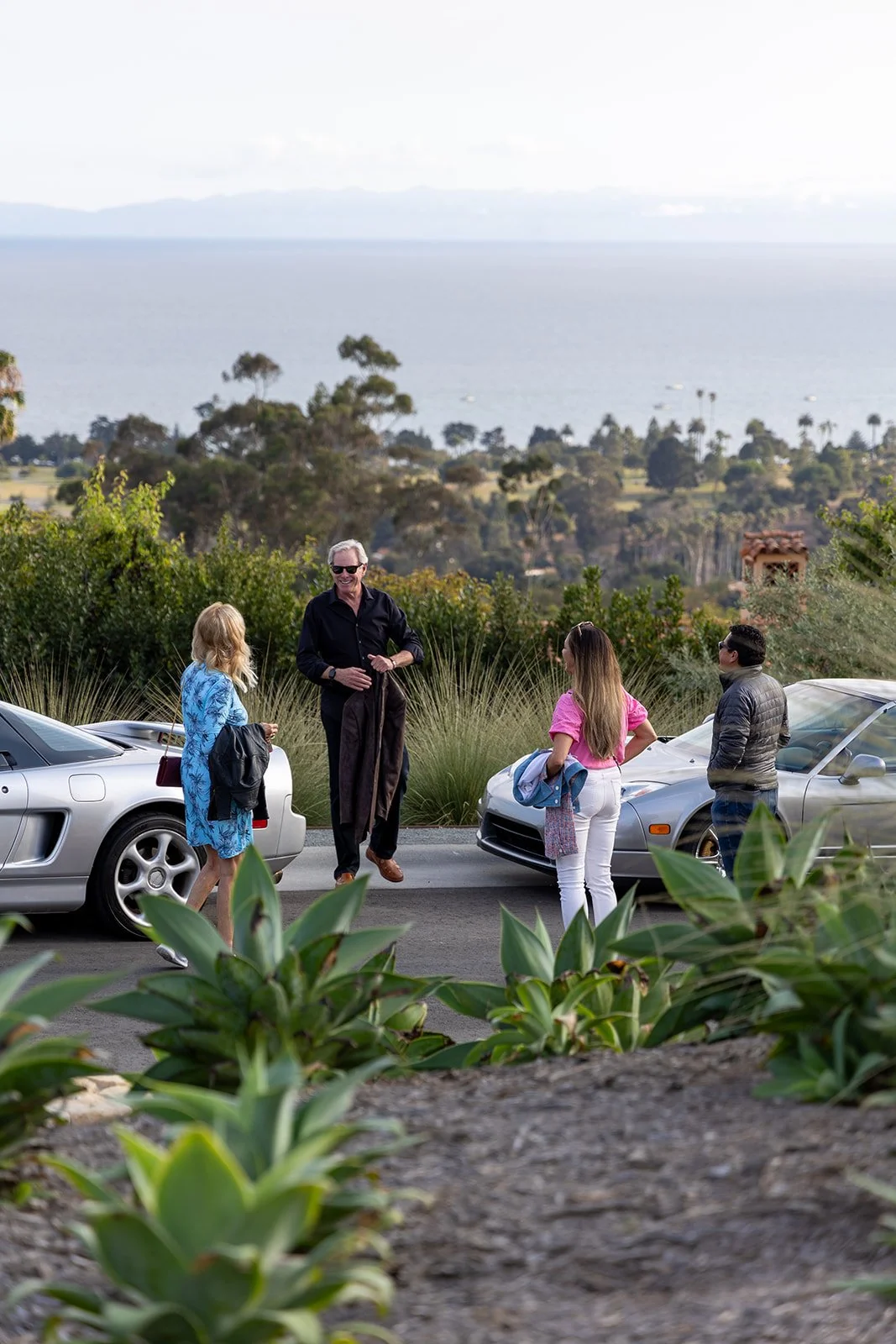 Four people standing on a paved road near parked cars, engaging in conversation, with a scenic background of trees, water, and mountains.