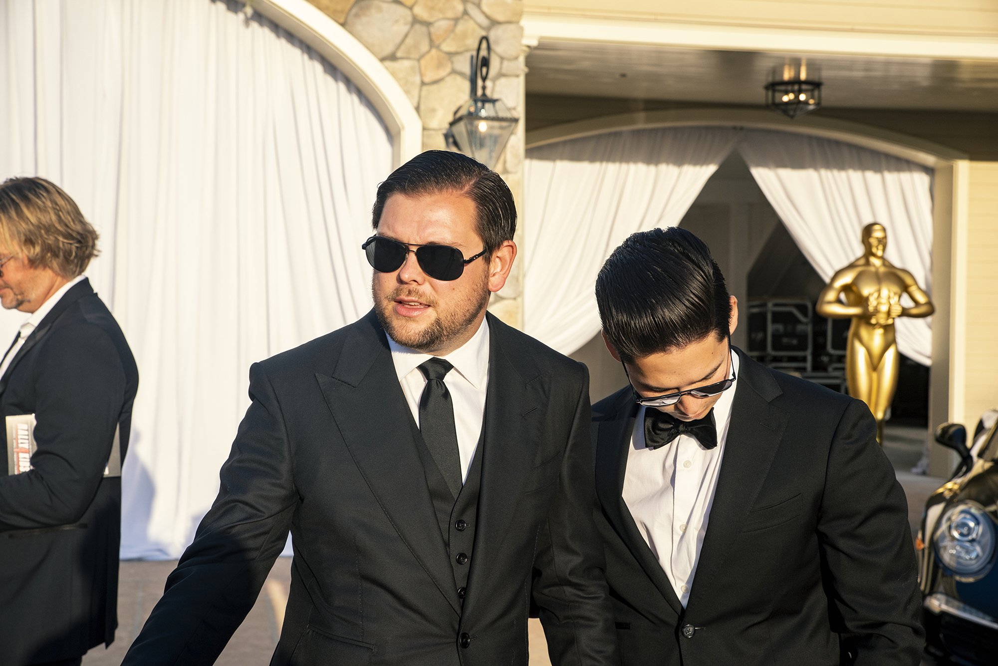 Two men in black tuxedos and sunglasses walking outdoors near a white tent, with a gold statue in the background.
