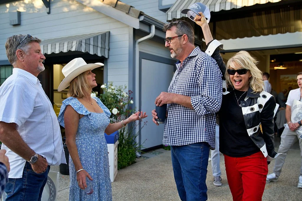 Four people engaged in conversation at an outdoor gathering, with two men and two women dressed casually, one woman wearing a sunhat and others wearing sunglasses.