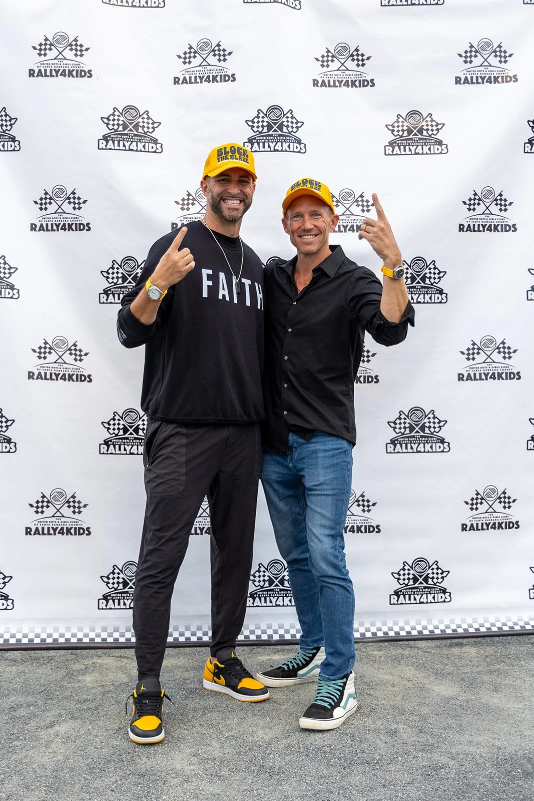 Two men standing together at a Rally4Kids event, posing for the photo. Both are pointing upward with their index fingers and are smiling. They are wearing yellow caps that say 'BLOCK THE BLAZE,' and casual clothing, including sneakers. The background