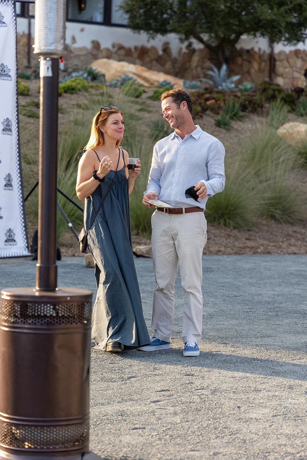 A woman and a man are standing outdoors, smiling and talking. The woman is holding a glass of wine, and the man is holding a booklet or brochure. There is a heater in the foreground, and the background features desert plants and a stone wall.