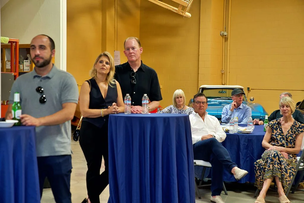 Group of people seated and standing around blue cocktail tables in a large indoor space with a yellow wall and a vintage blue car in the background