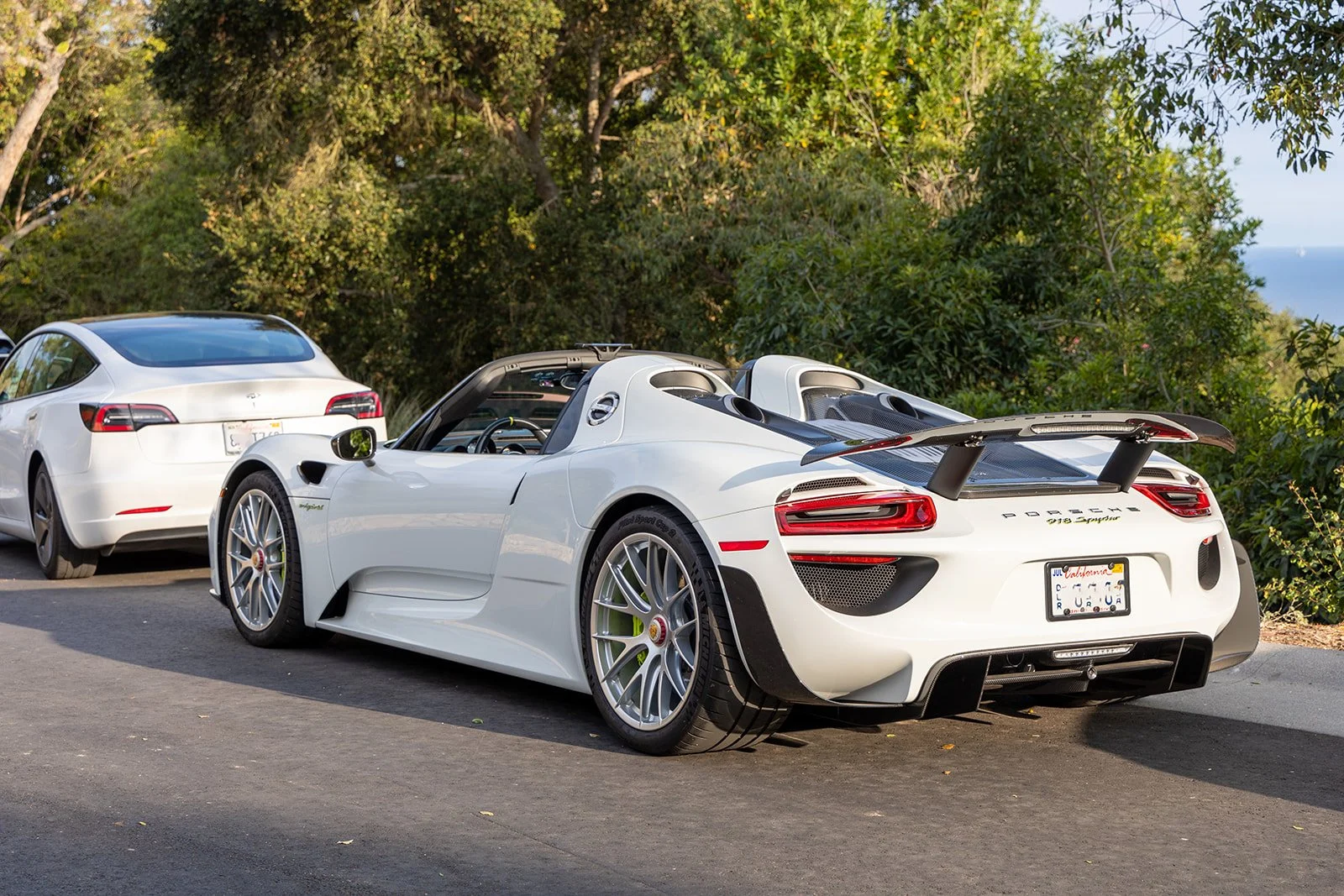 A white Porsche 918 Spyder sports car with a large rear wing, parked next to a white sedan in a parking lot surrounded by trees.