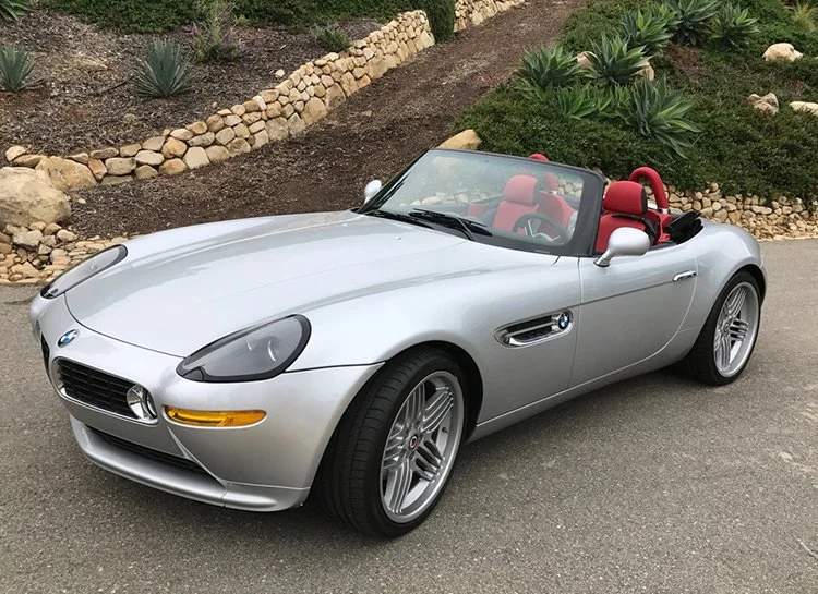 A silver BMW convertible sports car with red interior leather seats parked on a paved road, with a landscaped garden in the background.