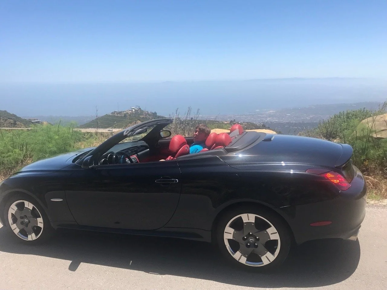 Black convertible car with red interior parked on a scenic hillside with ocean and land in the background.