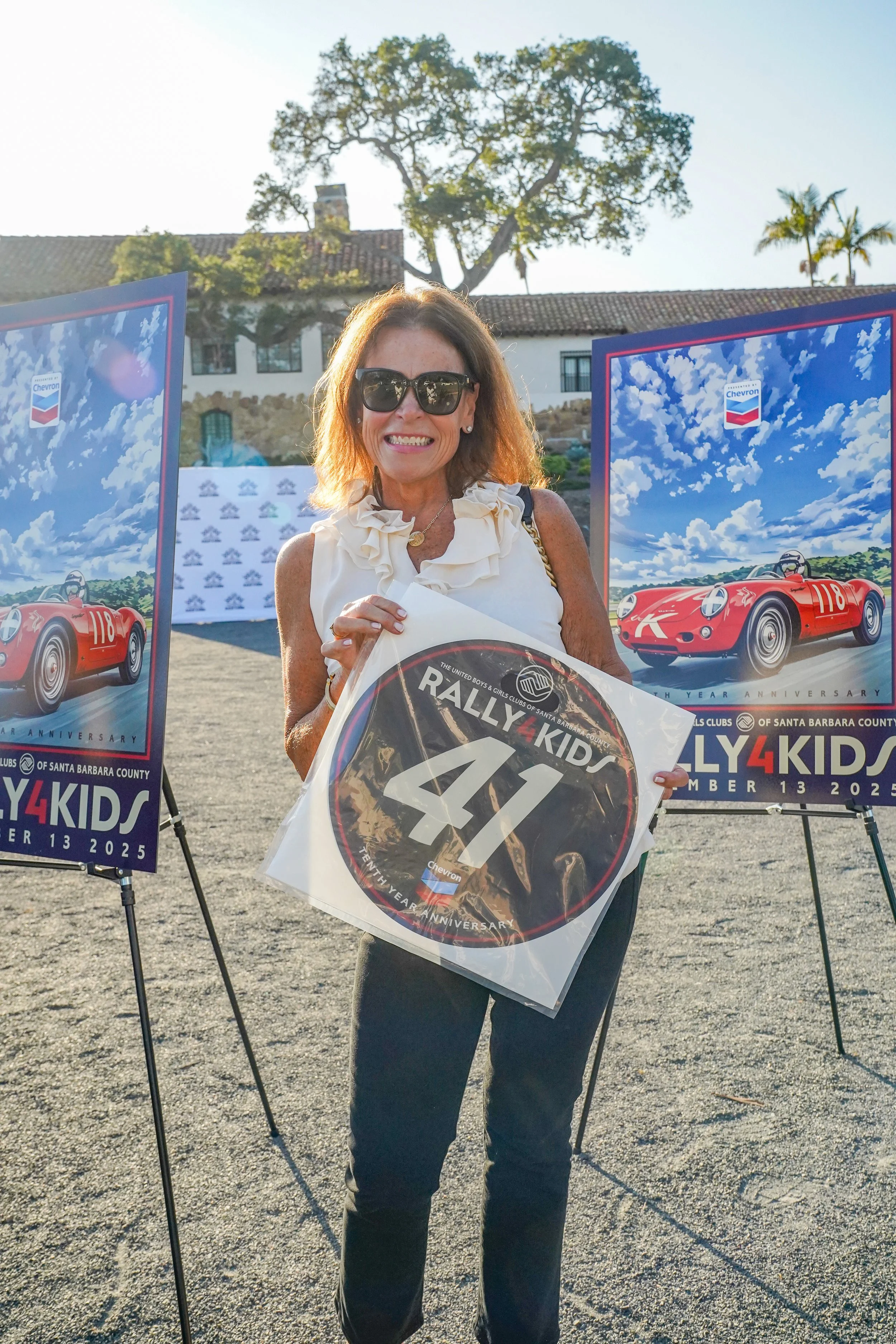 Woman holding Rally4Kids sign at event with vintage race car posters in background.