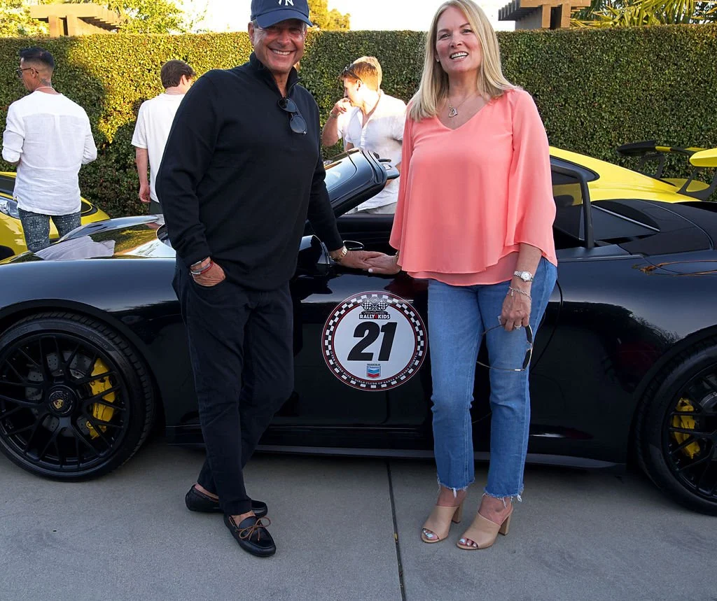 Two people, a man and a woman, stand next to a black sports car with a rally sticker number 21. The man is wearing a black outfit and a blue cap, smiling, and the woman is dressed in a peach-colored top and jeans, also smiling. In the background, thr