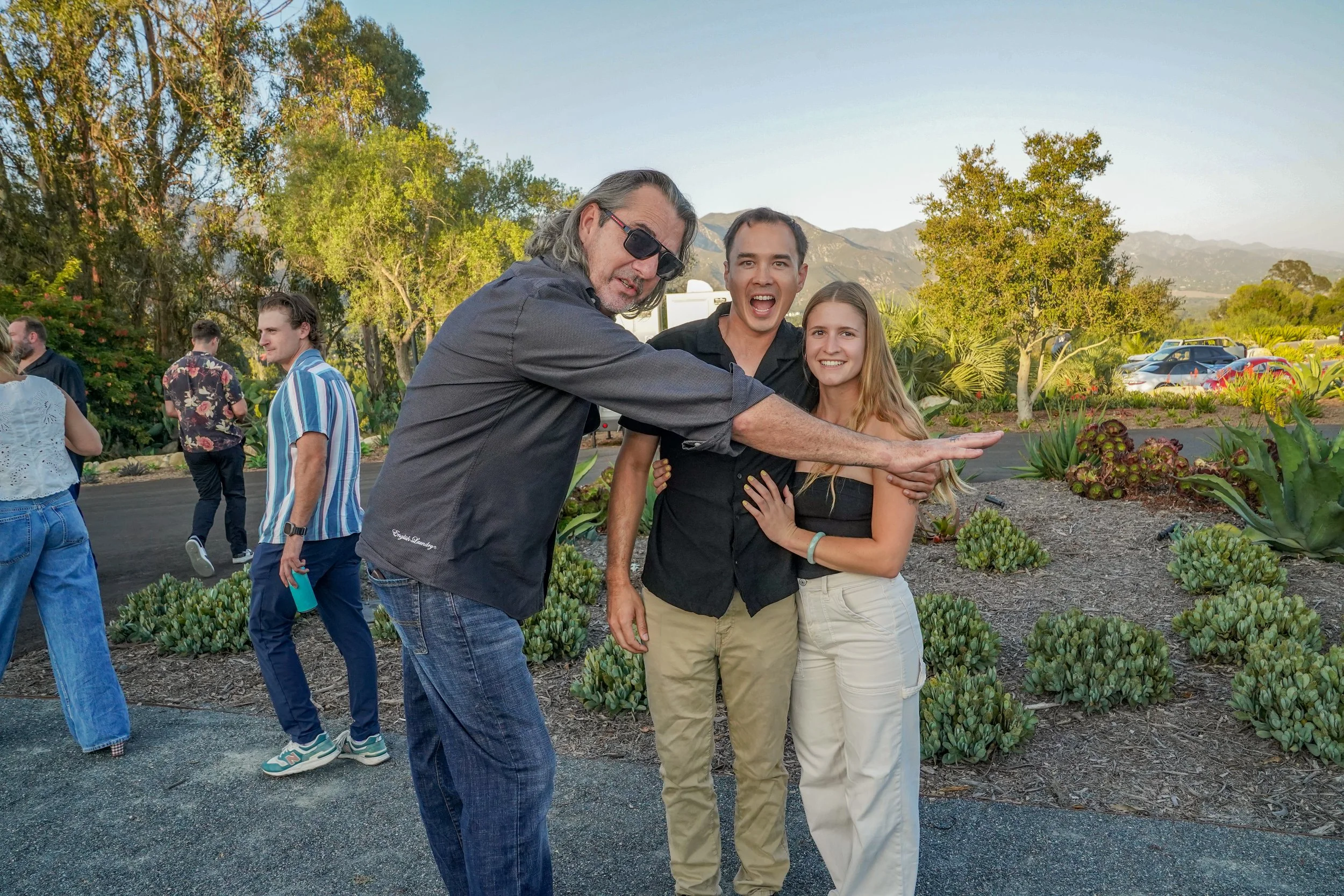A group of four people, three men and one woman, posing outdoors in a garden with trees and mountains in the background. One man is extending his arm sideways, with the others smiling or reacting around him.