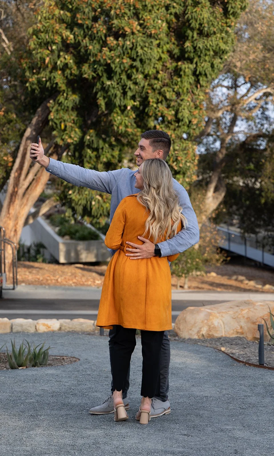 A smiling couple takes a selfie outdoors, with the man’s arm extended holding a smartphone. The woman has long blonde hair and wears a mustard yellow coat, while the man wears a light gray shirt and dark pants. They are standing on a gravel surface w