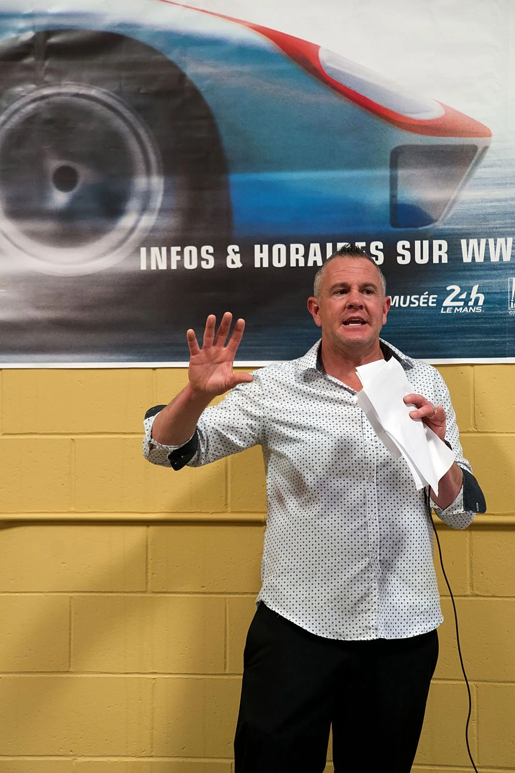 Man giving a presentation or speech, holding papers, standing in front of a large banner with race car graphic, yellow wall background.
