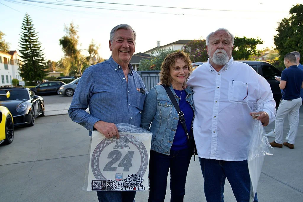Three people standing together outdoors at a car gathering, smiling, with parked cars and trees in the background.