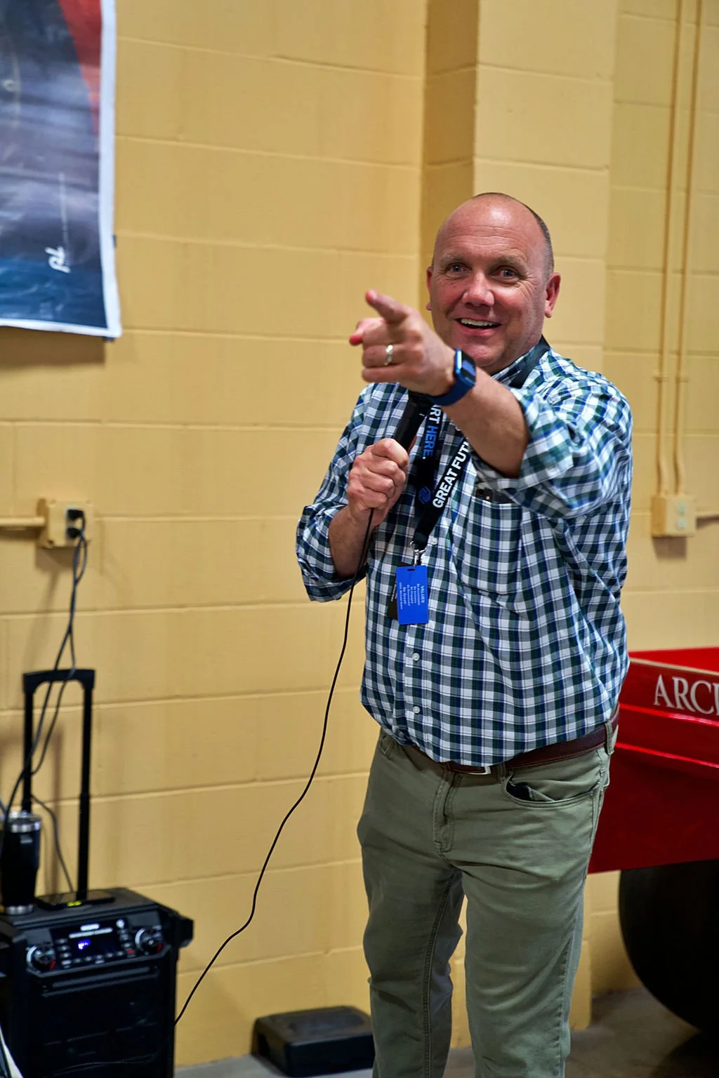 A man with a shaved head wearing a checkered shirt holding a microphone, pointing towards the camera, standing in front of a yellow wall with a poster on it.