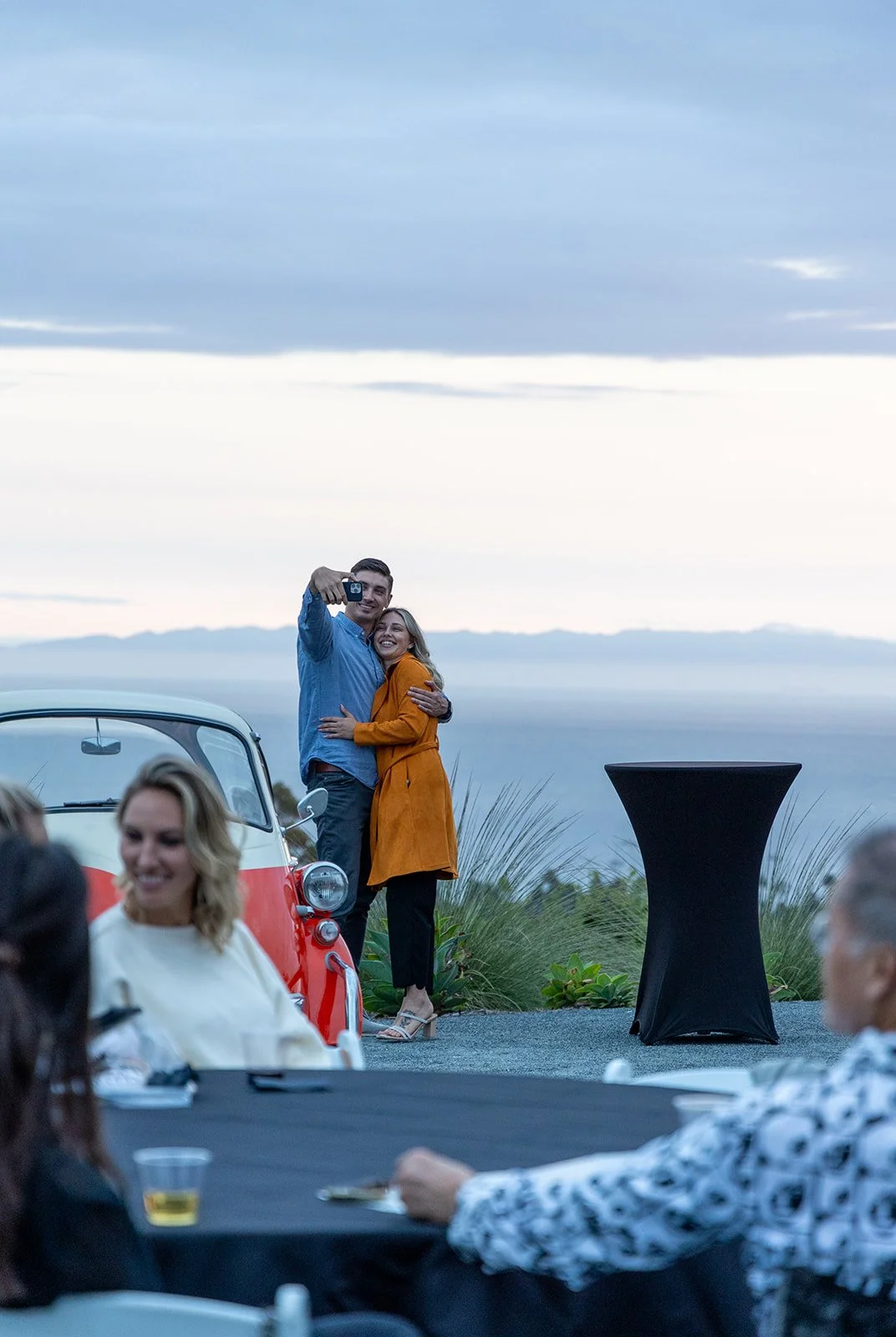 A couple takes a selfie at an outdoor event during sunset, with a vintage car and a view of the water in the background, while other guests sit at a table.