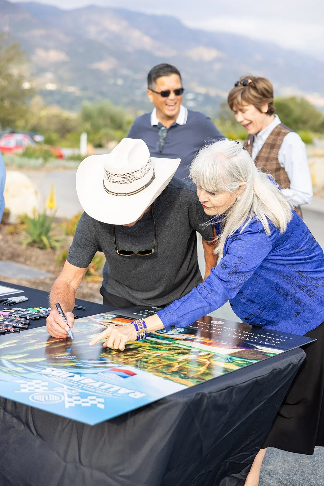 A man in a black shirt and white cowboy hat signs a colorful poster at an outdoor event, with a woman in a blue jacket pointing at the poster and people in the background laughing and talking.