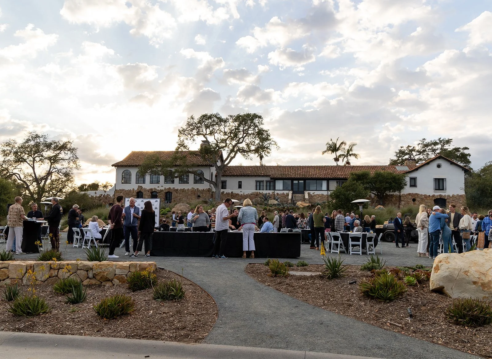 People gathering at an outdoor event on the grounds of a large white house with a red tile roof, surrounded by trees and landscaping, under a partly cloudy sky.