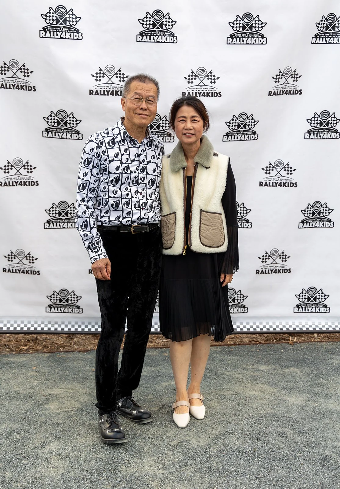 A man and a woman stand together in front of a branded backdrop with logos for Rally4Kids, smiling at the camera. The man is wearing a patterned shirt, black pants, and shiny black shoes. The woman is wearing a black pleated skirt, white shoes, and a