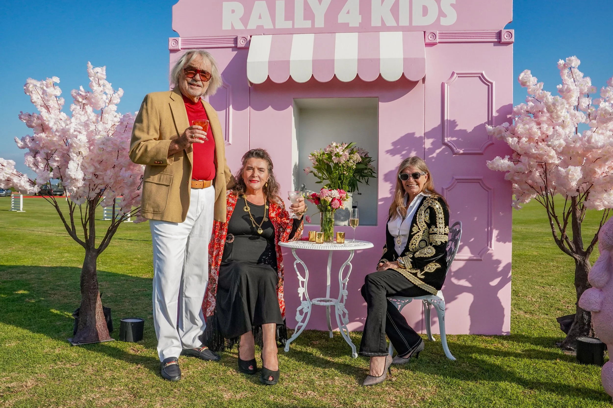 Three people, two women and one man, celebrating outside in front of a pink backdrop with the sign 'Rally 4 Kids'. The man is standing, dressed in a tan jacket, red shirt, white pants, and sunglasses, holding a glass. The woman on the left is seated,