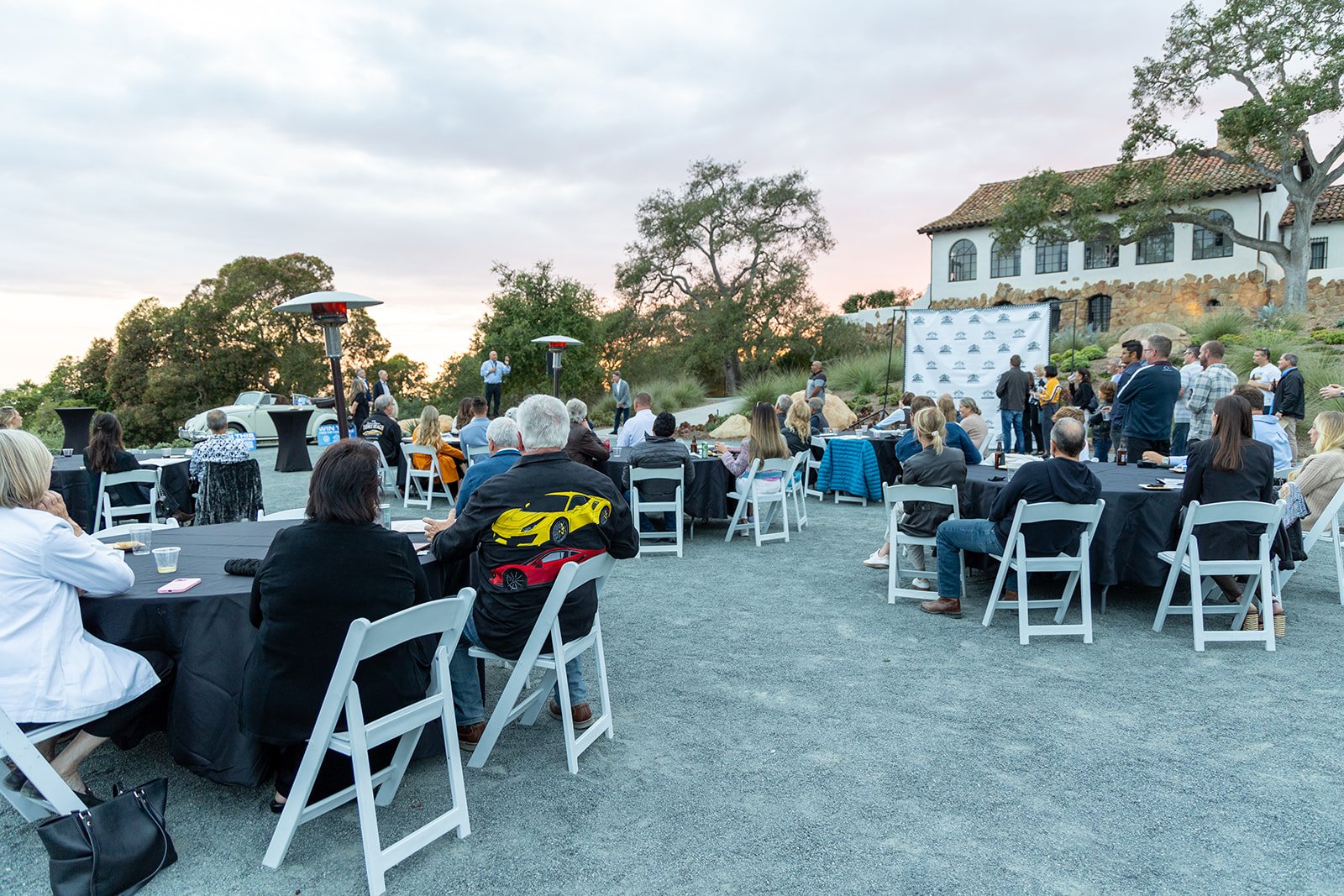 Outdoor business event or conference with attendees seated at tables, a speaker on stage, and a backdrop with logos, during sunset at a scenic venue.