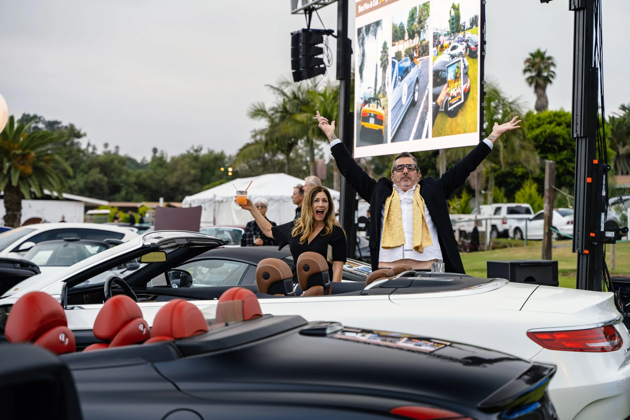 Two people celebrating in a convertible with palm trees and parked cars in the background. One person is holding a drink and smiling, while the other is standing with arms raised next to a large screen showing images of cars and other objects.