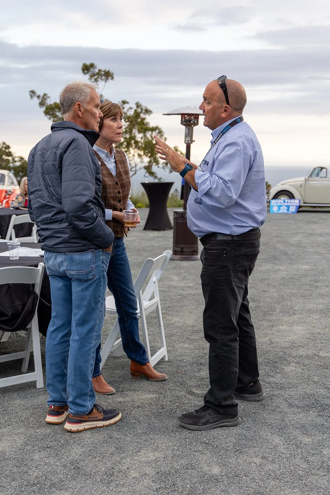 Three people standing outdoors having a conversation during an event, with tables and vintage cars in the background.