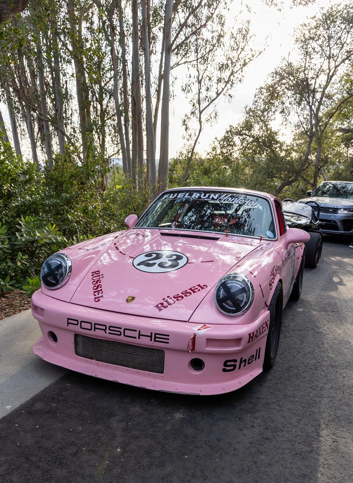 A vintage pink Porsche race car with the number 23 on the hood, surrounded by trees and other cars in the background at a gathering or event.