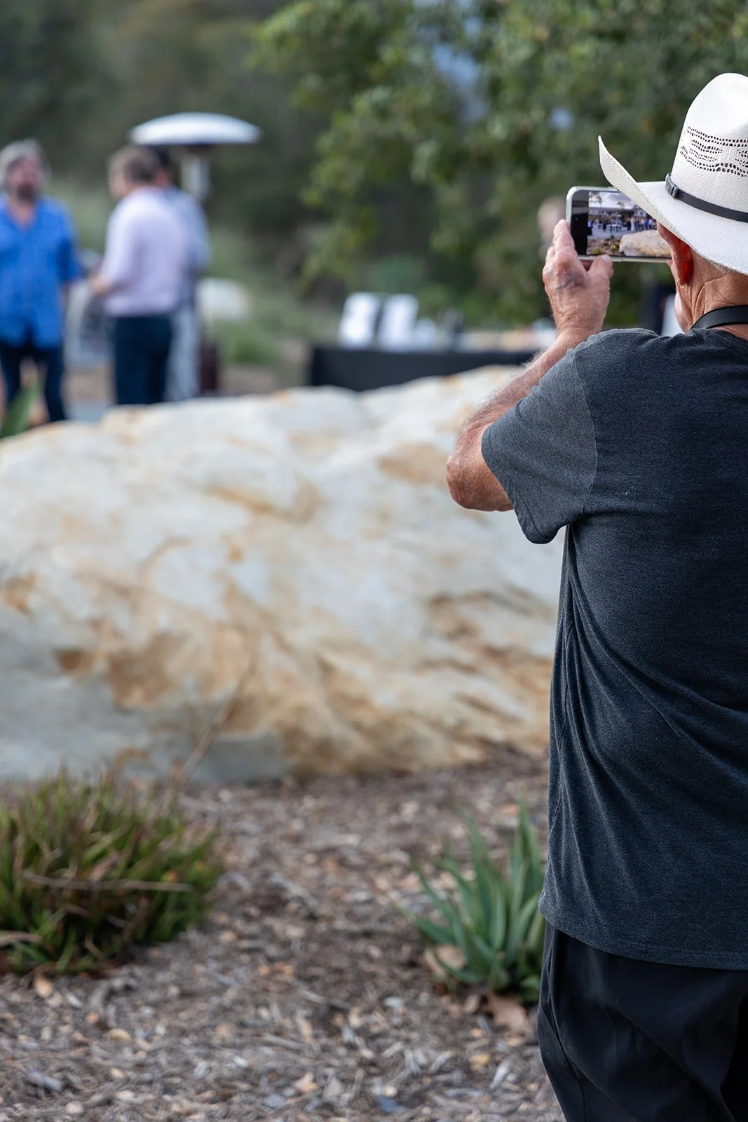 An elderly man wearing a white hat and black t-shirt is taking a photo of a group of three people standing near a large rock using his smartphone. The background includes trees and outdoor setting.