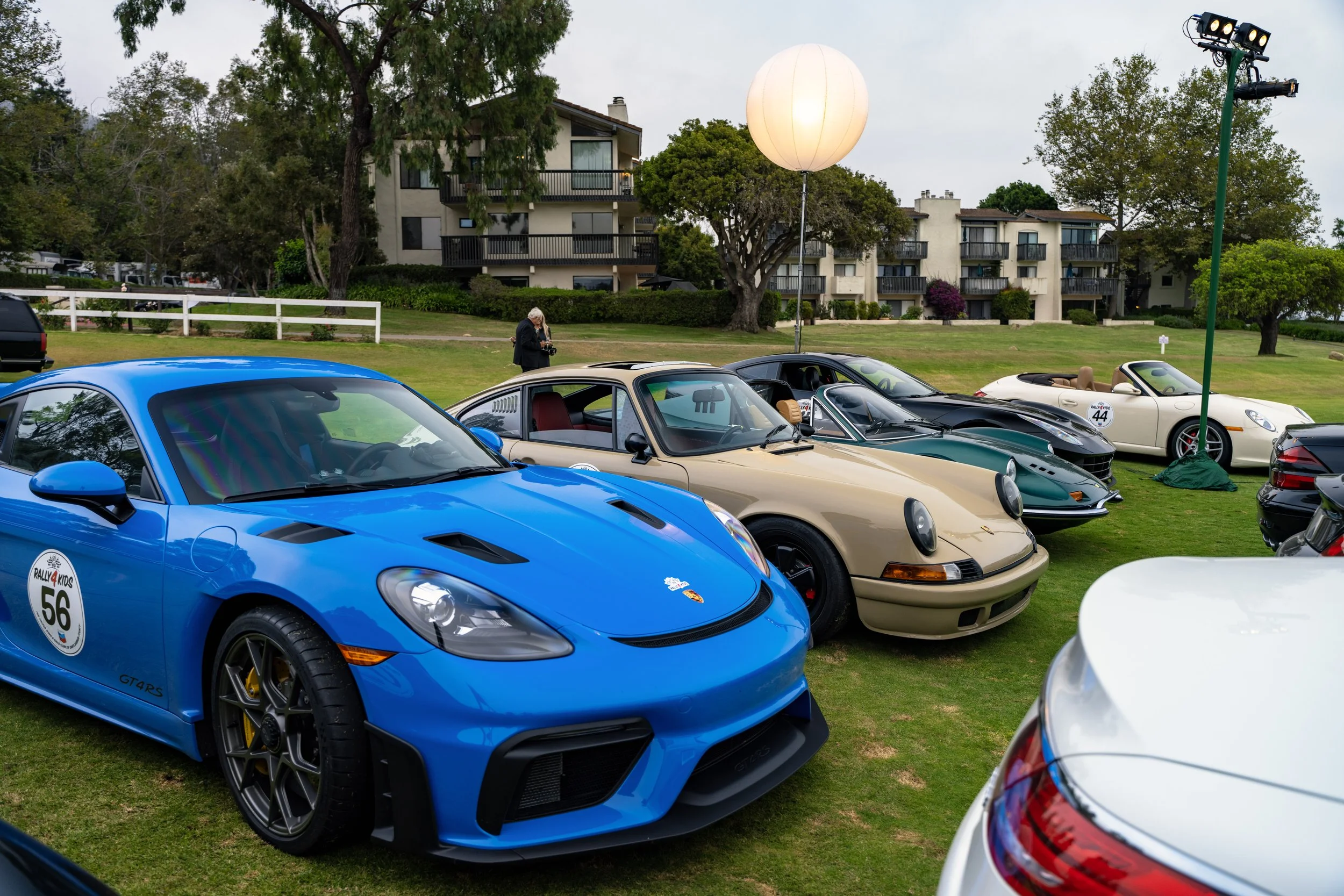 Multiple sports cars, including a blue Porsche 718 Cayman and a beige Porsche 911, parked on a grassy area at a car show with a large white balloon and spotlights in the background.