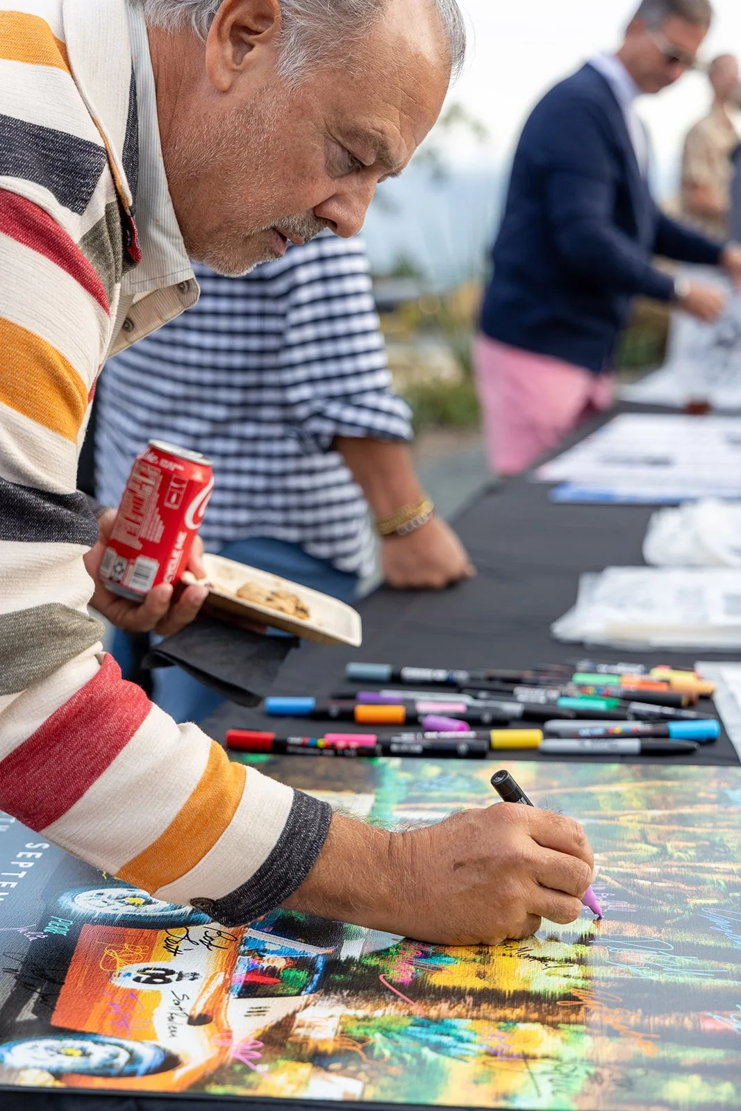 A man in a striped sweater signing a colorful poster at an outdoor event, with markers and other people in the background.