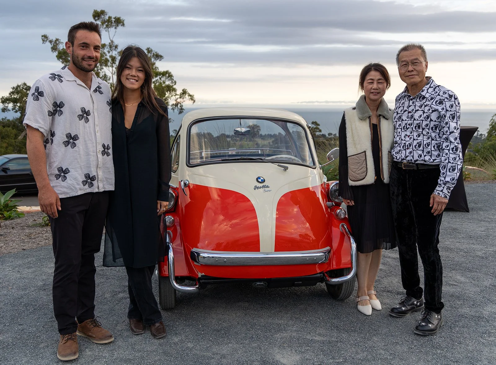 Five people standing outdoors in front of a vintage BMW Isetta car with a scenic background of trees and hills, during a cloudy day.