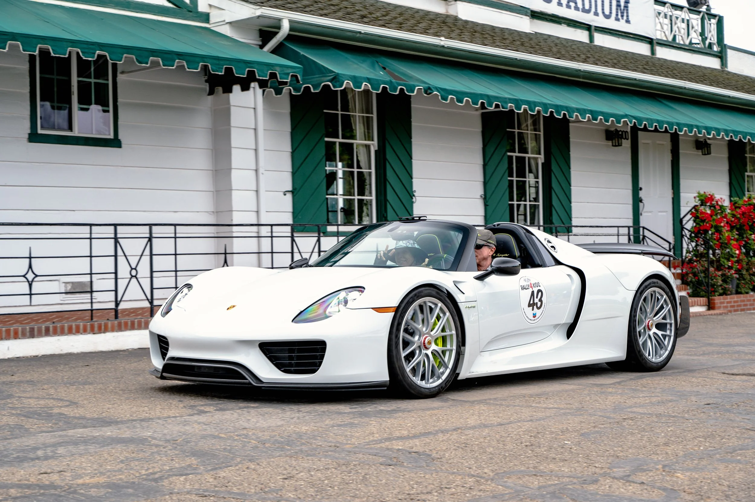 A white Porsche sports car with a race number 43 parked in front of a white building with green shutters and an awning.
