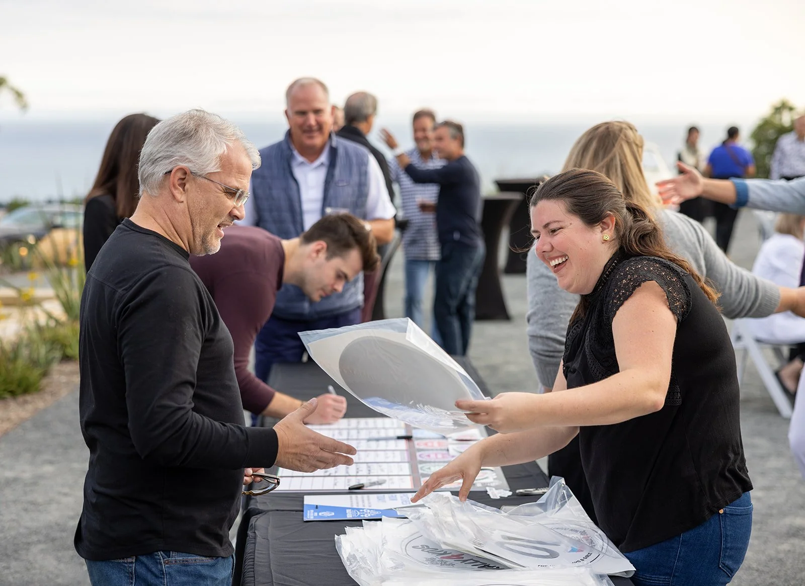 People attending an outdoor event, with individuals at a registration or check-in table, exchanging documents, smiling, and engaging in conversation near a body of water.