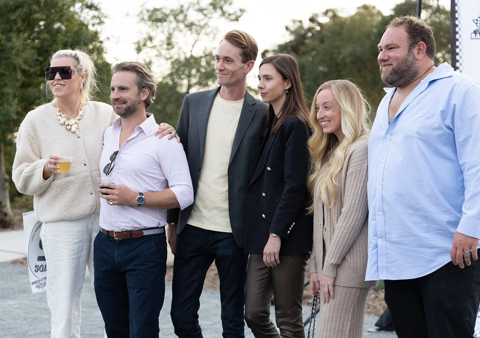 Group of six people standing outdoors, smiling, with trees in the background. Some holding drinks, dressed in casual and business casual attire.