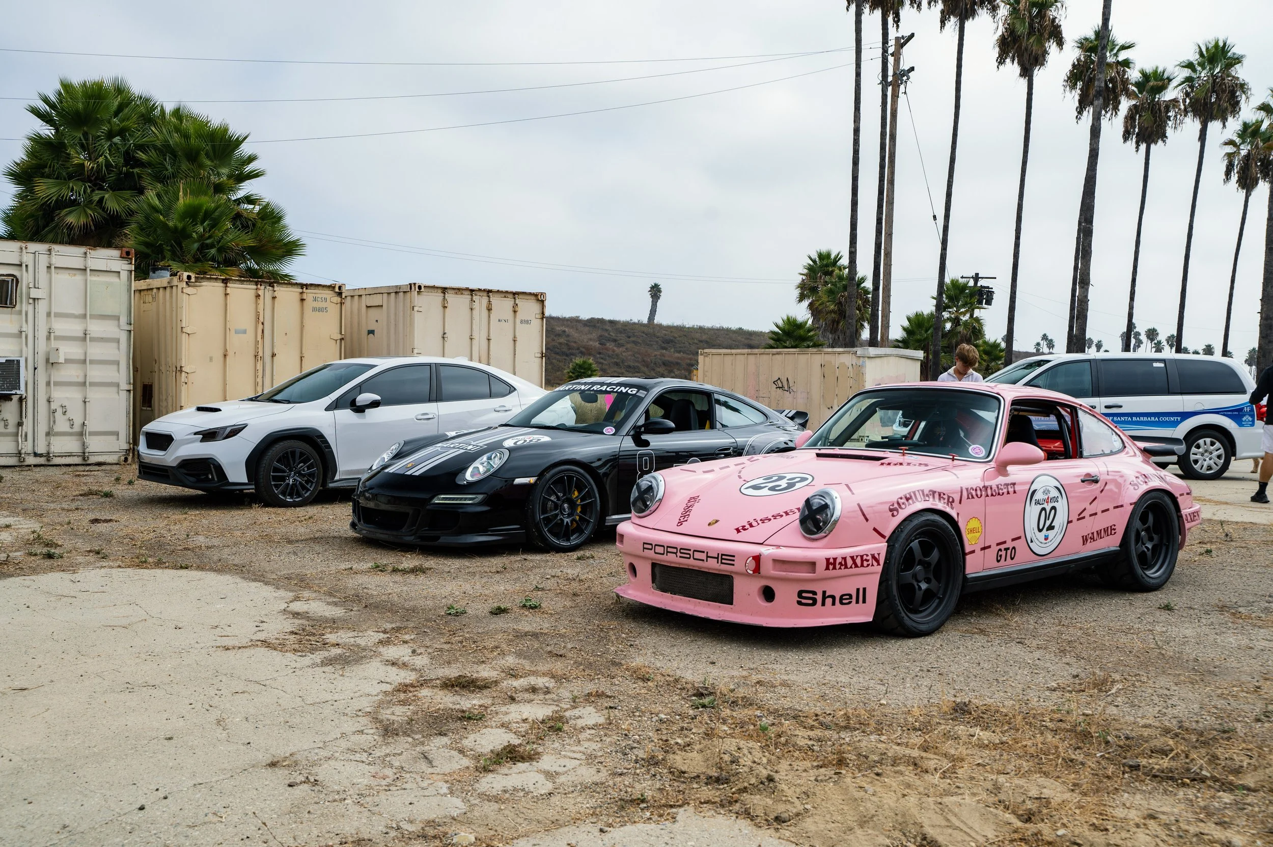 A lineup of three sports cars parked on a dirt lot, including a pink Porsche with vintage racing decals, a black Porsche 911, and a white Subaru. There are palm trees, shipping containers, and a police SUV in the background.