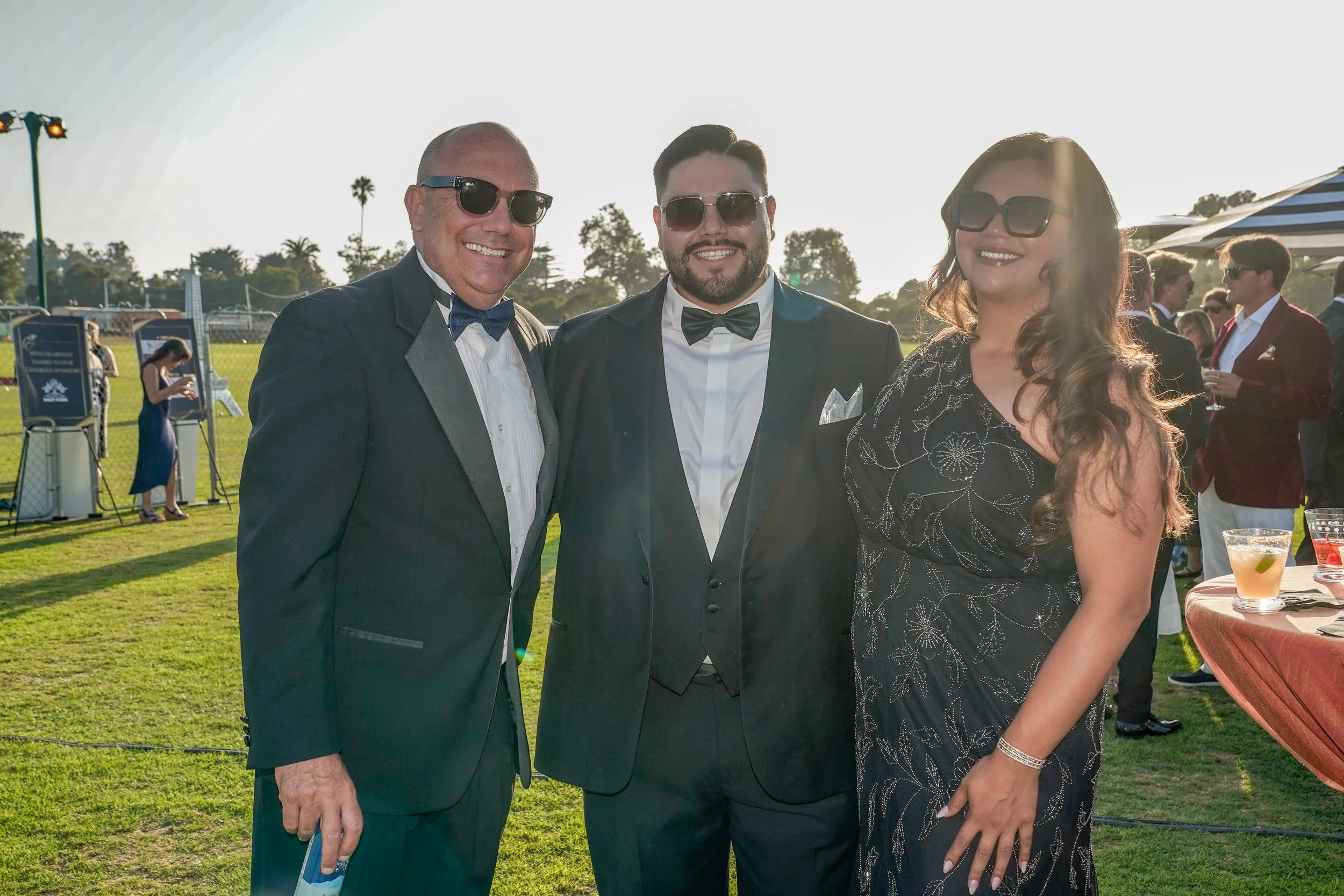 Three people in formal tuxedos and dress at an outdoor event, wearing sunglasses, smiling for the camera, with a grassy area and other guests in the background