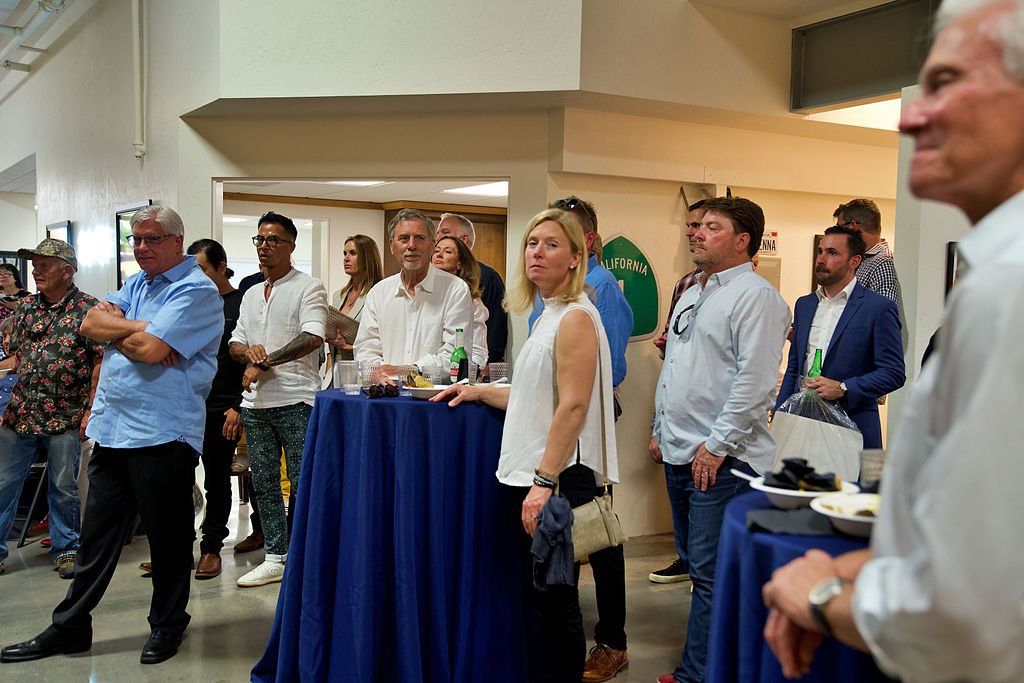 Group of people at a social gathering or reception, some standing, some holding drinks, in a room with beige walls and a blue tablecloth.