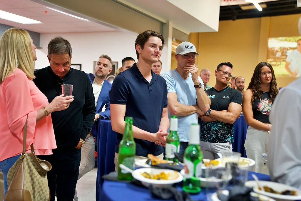 Group of people standing around a table with food and drinks, listening to a woman speak.