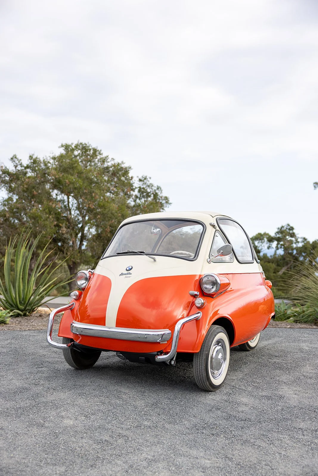 A vintage BMW Isetta microcar in orange and cream parked on a gravel surface with desert plants and trees in the background.