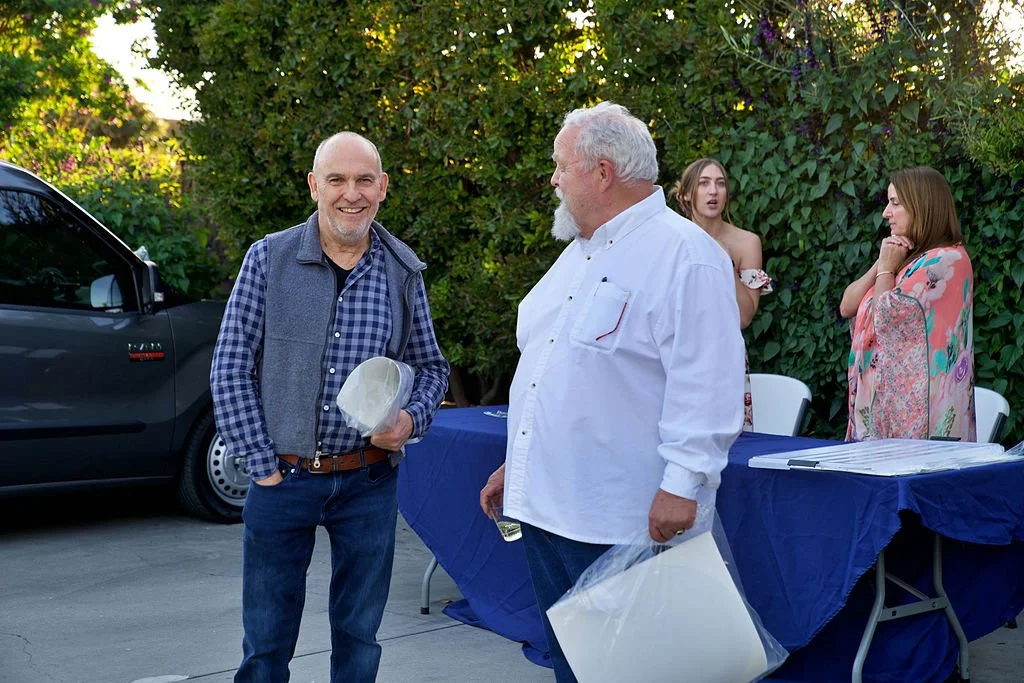 Two older men talking and smiling at an outdoor gathering, with women in the background and a car parked nearby.