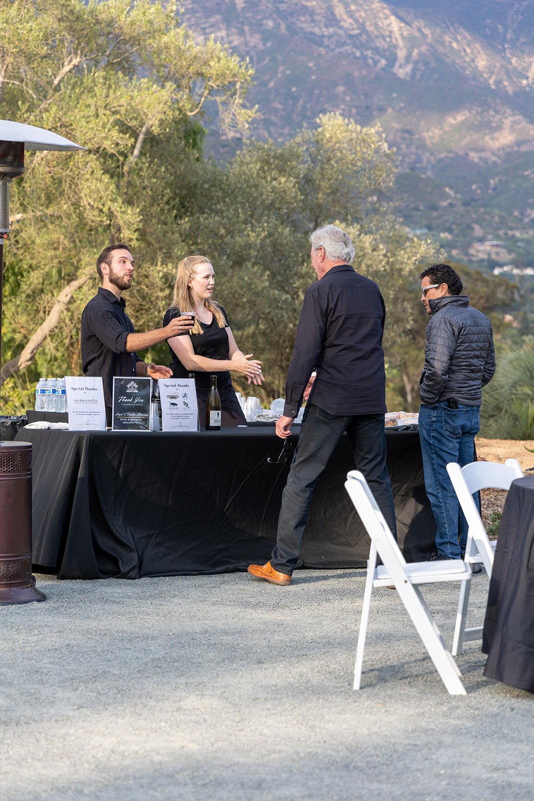 People attending an outdoor event with food and drinks, set against a scenic mountain backdrop, and a table with signs and bottles.