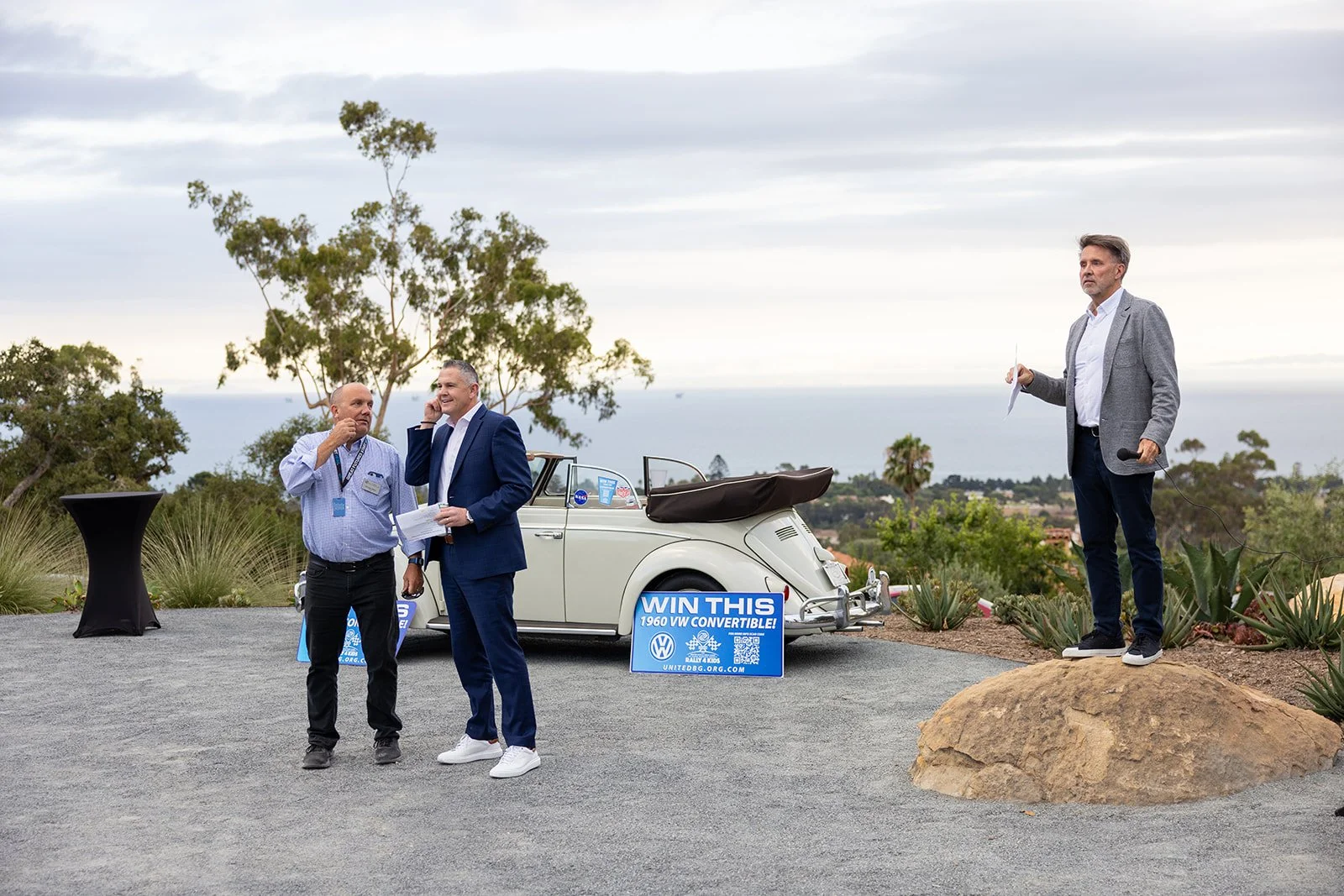 Two men chatting, one in a suit and the other in a striped shirt, beside a classic cream-colored convertible car with a 'Win This' sign, outdoors with cloudy sky and trees in the background. A man holding a microphone stands on a rock nearby.